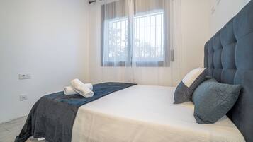 Un dormitorio con cama, ventana con cortinas translúcidas y una toalla sobre la cama.
