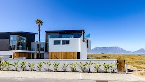 A modern two-story building with large windows, a wooden fence, and a view of mountains in the background. - Ocean 9 - Pool-Suite (Cape Town)