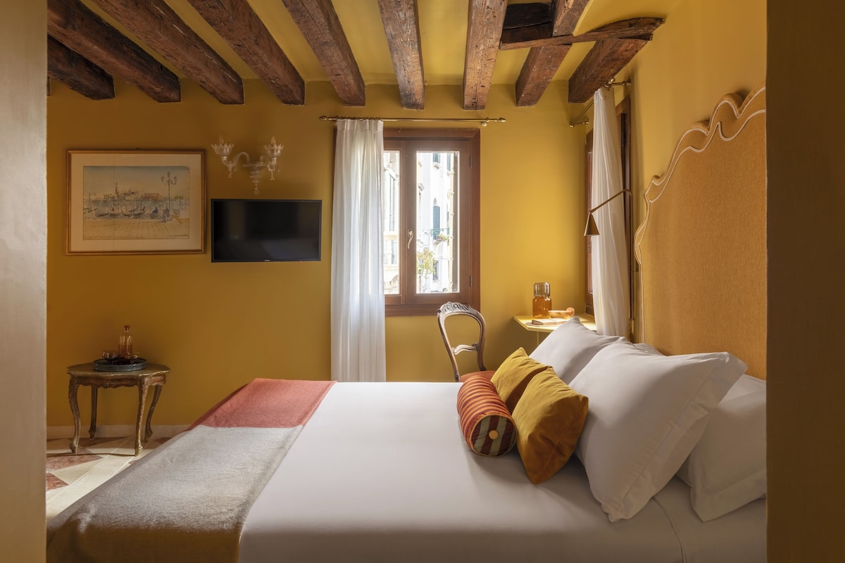Una habitación de hotel con una cama, televisor, ventana con cortinas, una mesita con una vela y un cuadro en la pared.