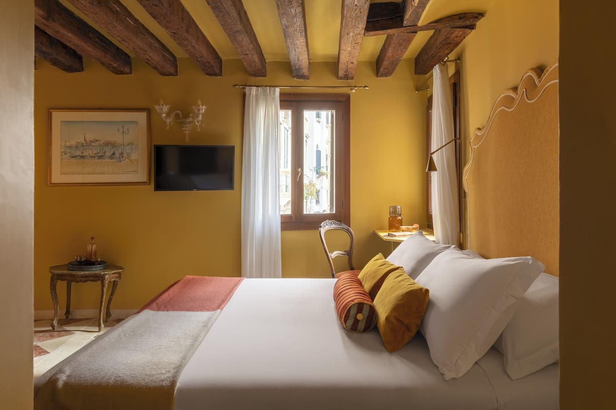 A hotel room with a bed, a television, a window with curtains, a small table with a candle, and a painting on the wall.