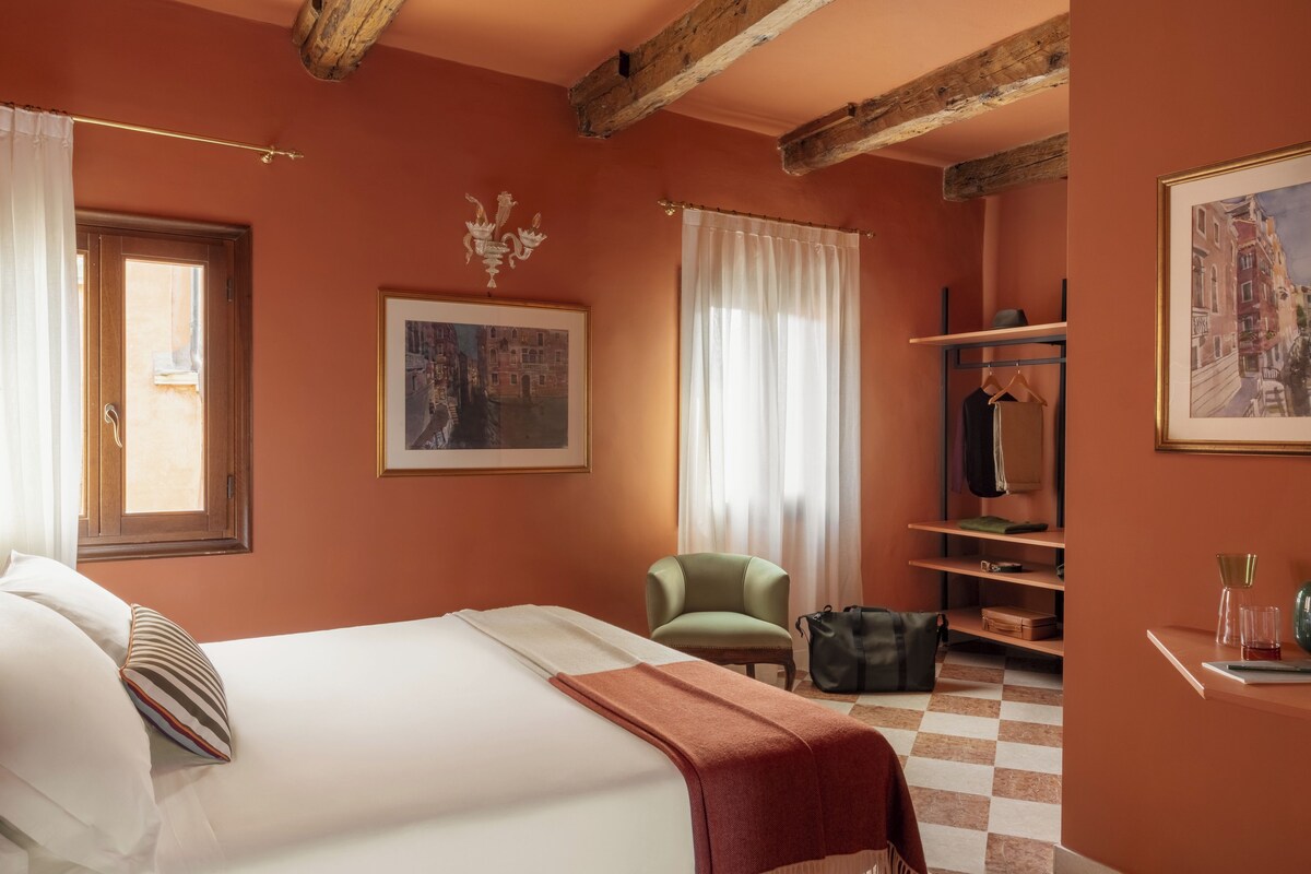 Une chambre d’hôtel avec un lit, une chaise, un bureau, un porte-manteau et une fenêtre avec des rideaux.