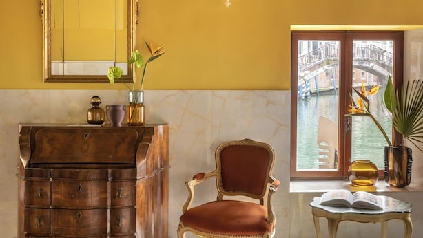 Une pièce avec un meuble en bois, une chaise, une table sur laquelle se trouve un livre, une fenêtre donnant sur un canal et un tapis à motifs.