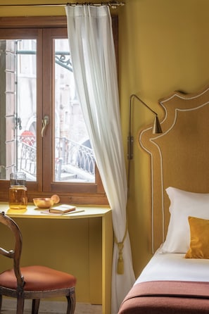 Ein Hotelzimmer mit einem Holztisch, einem Stuhl, einem Bett mit weißem und orangefarbenem Kopfkissen und Blick auf die Straße durch ein Fenster mit Vorhang.