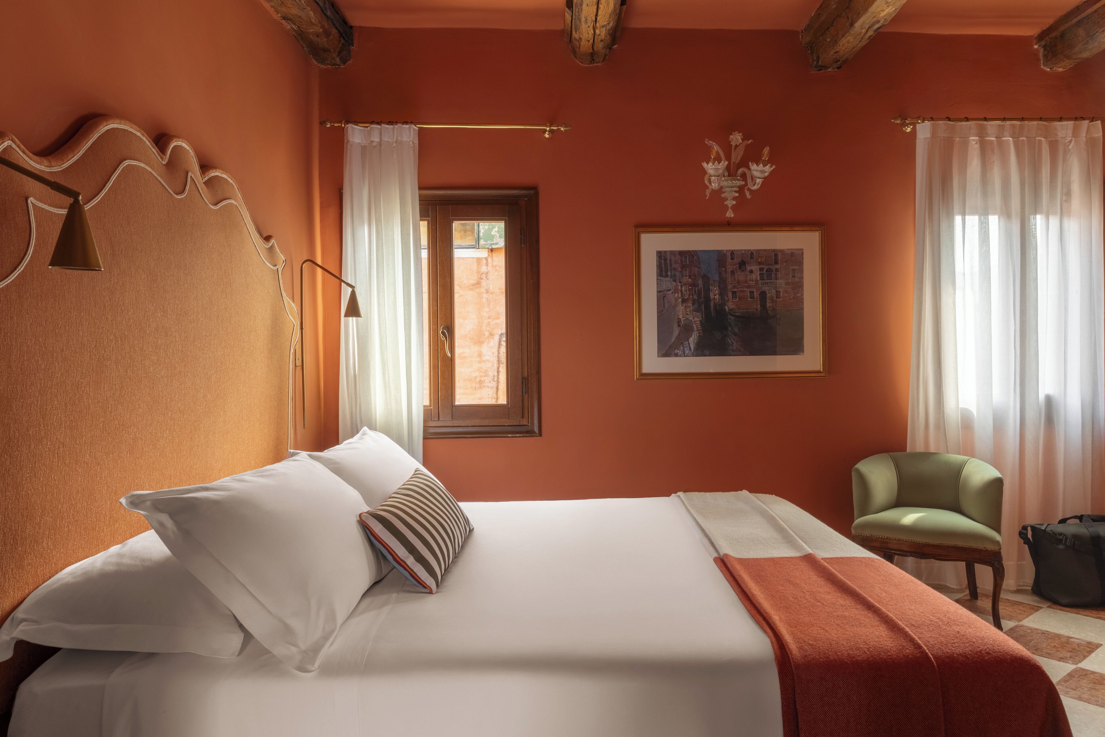 A hotel room with a large bed, a chair, a window with curtains, and a painting on the wall.