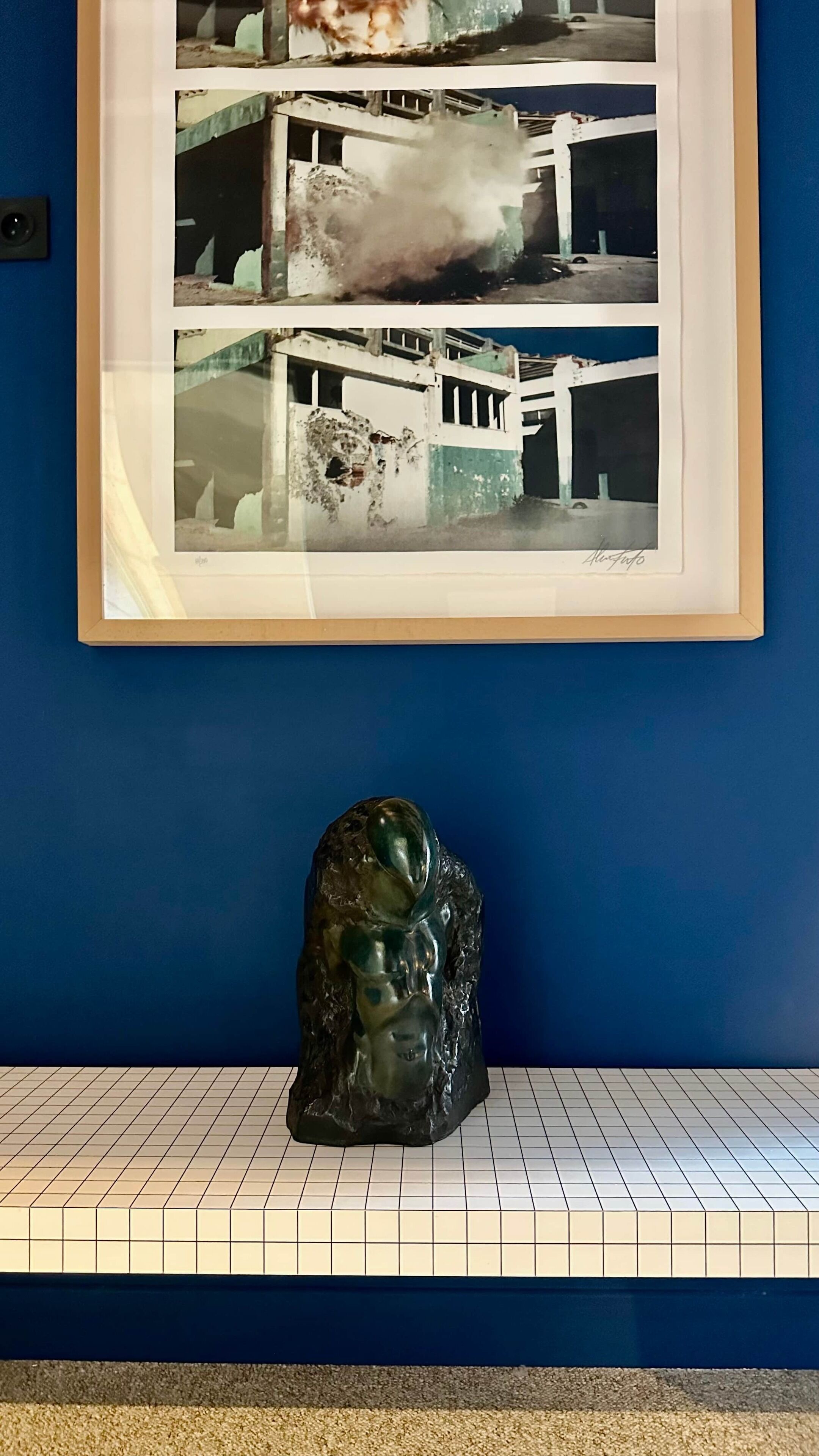 Een ingelijste foto van een vervallen gebouw op een blauwe muur, met een sculptuur eronder.