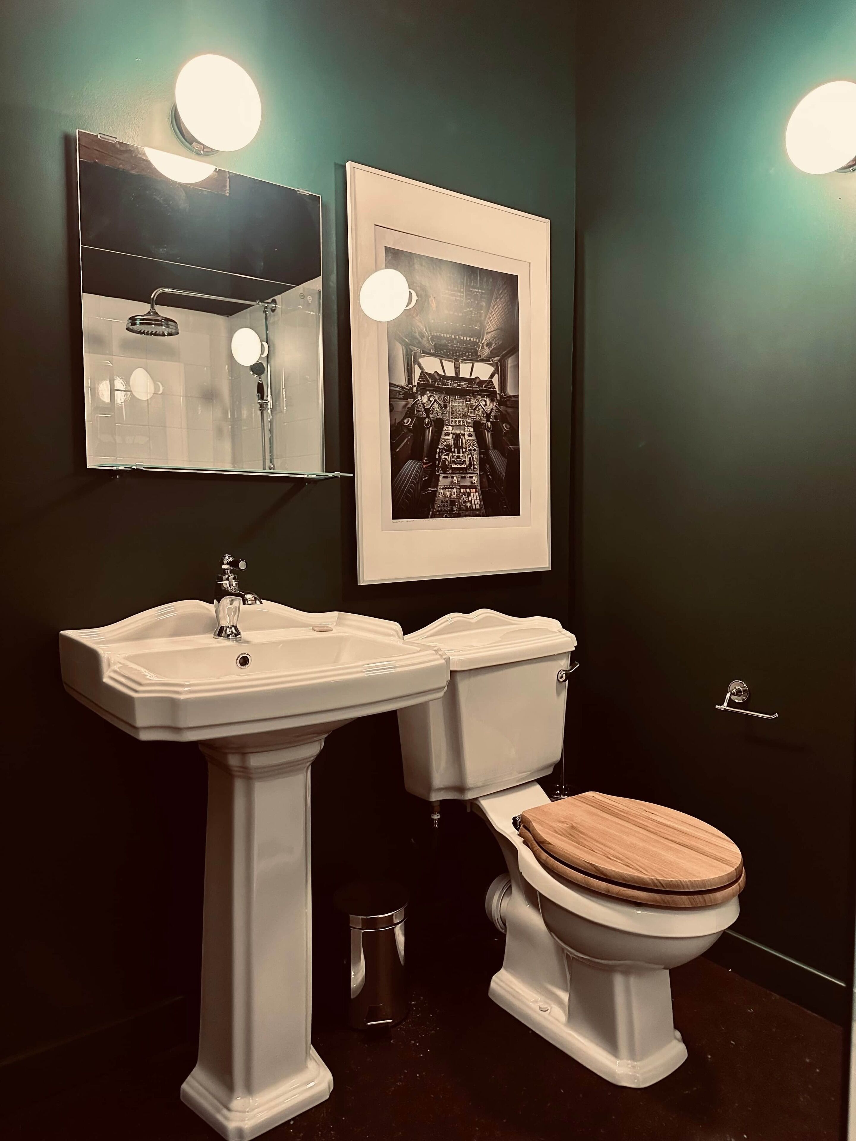 A bathroom with a pedestal sink, a toilet with a wooden seat, a framed picture on the wall, and two wall-mounted lights.