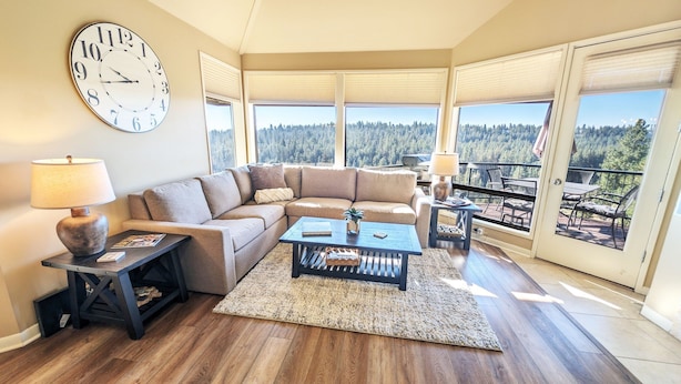 Ein Wohnzimmer mit einer großen Uhr, einem Sofa, einem Couchtisch und Blick auf einen Wald durch große Fenster.