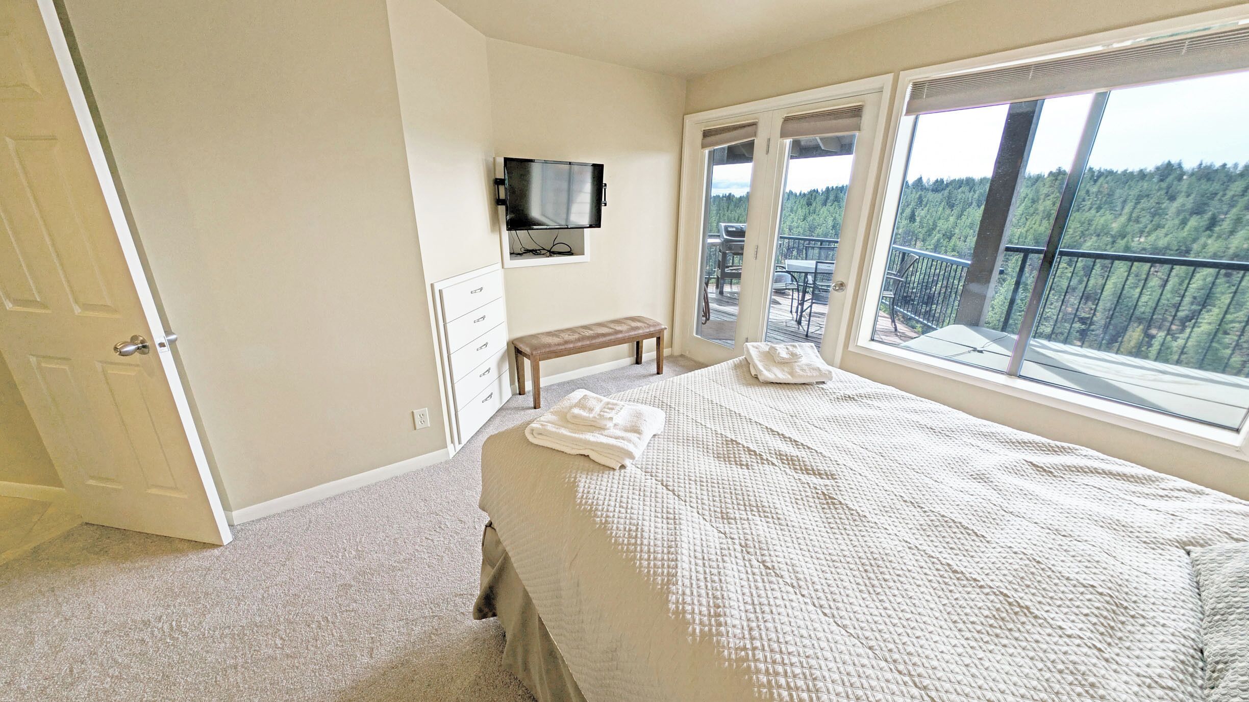 A bedroom with a large bed, a bench, a TV mounted on the wall, and a balcony with a view of trees.