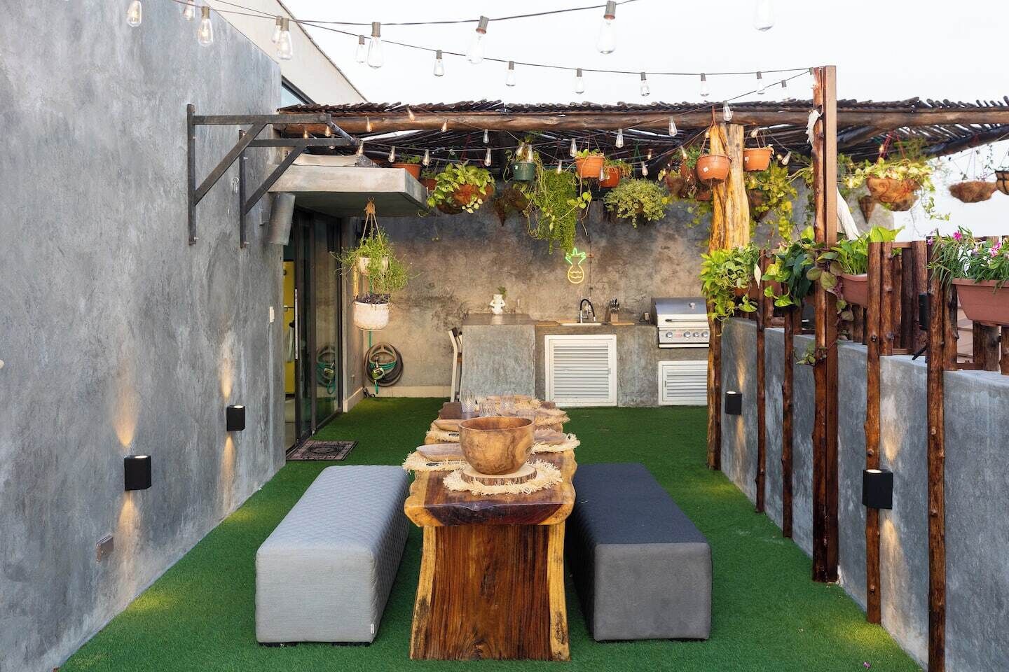 Un espacio exterior pequeño con césped artificial, una mesa de madera, dos bancos grises, macetas colgantes y una parrilla.