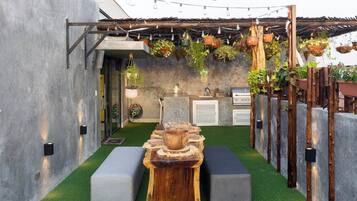Un espacio exterior pequeño con césped artificial, una mesa de madera, dos bancos grises, macetas colgantes y una parrilla.