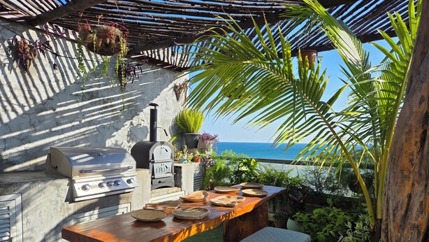 An outdoor kitchen with a grill, a table set with plates, and a view of the ocean.