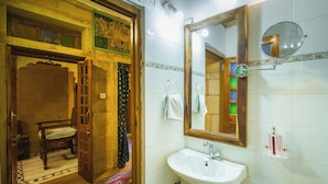 A bathroom with a wooden door, a white sink, a mirror, and a tiled wall. - Hotel Rajmandir  (Jaisalmer)