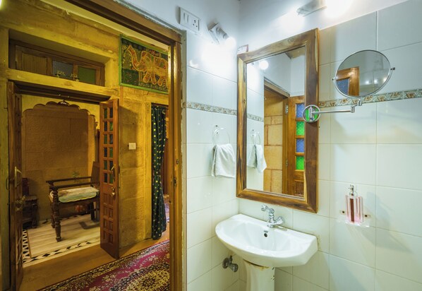 A bathroom with a wooden door, a white sink, a mirror, and a tiled wall. - Hotel Rajmandir  (Jaisalmer)