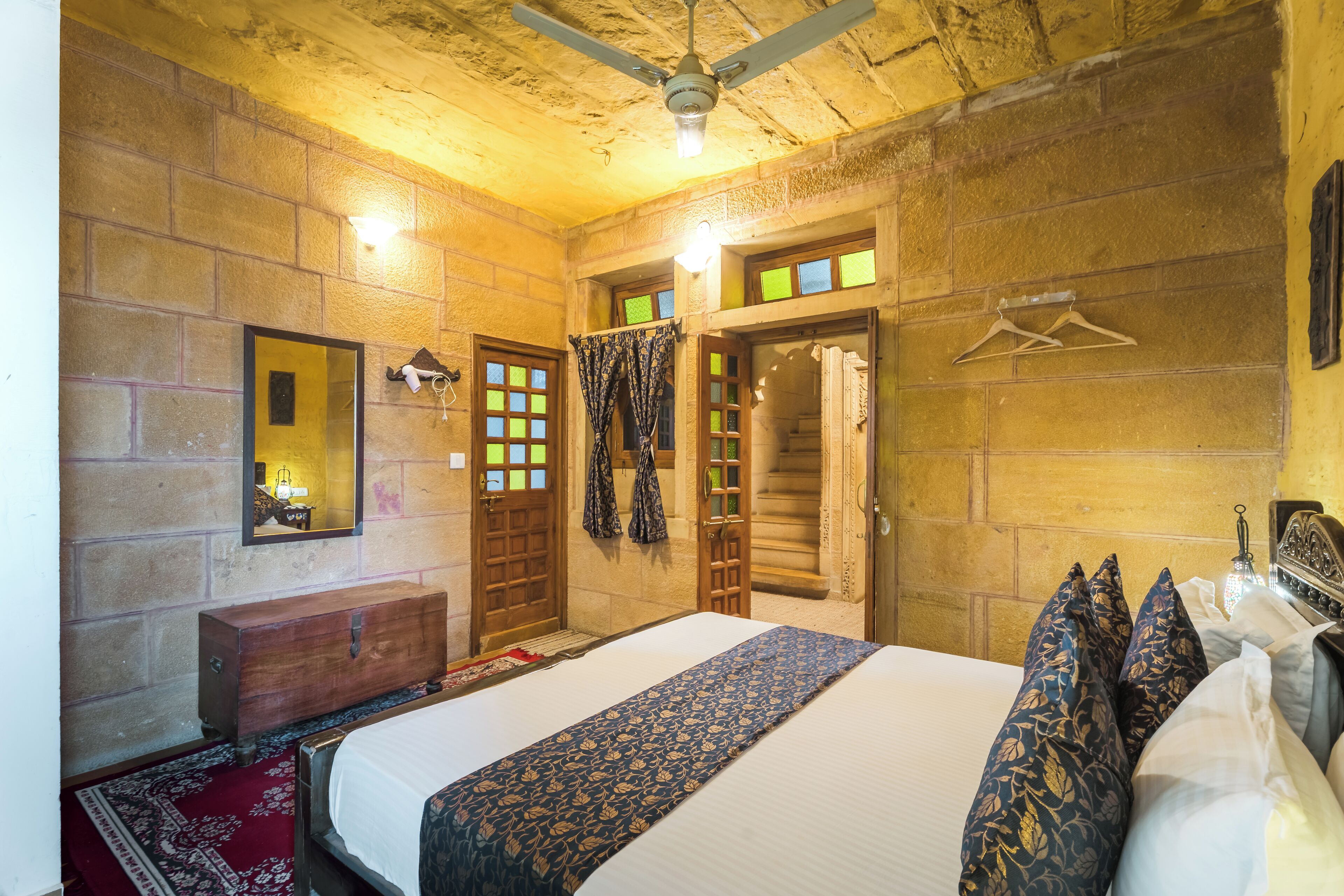 A hotel room with a large bed, a wooden dresser, a mirror, a ceiling fan, and a view of the outside through the window.