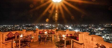 Un área de comedor al aire libre con mesas y sillas, una lámpara central grande y vistas de la ciudad por la noche.