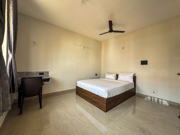 A bedroom with a bed, a desk, a chair, a ceiling fan, and a light switch on the wall. - RK Homes Service Apartments (Vasco da Gama)