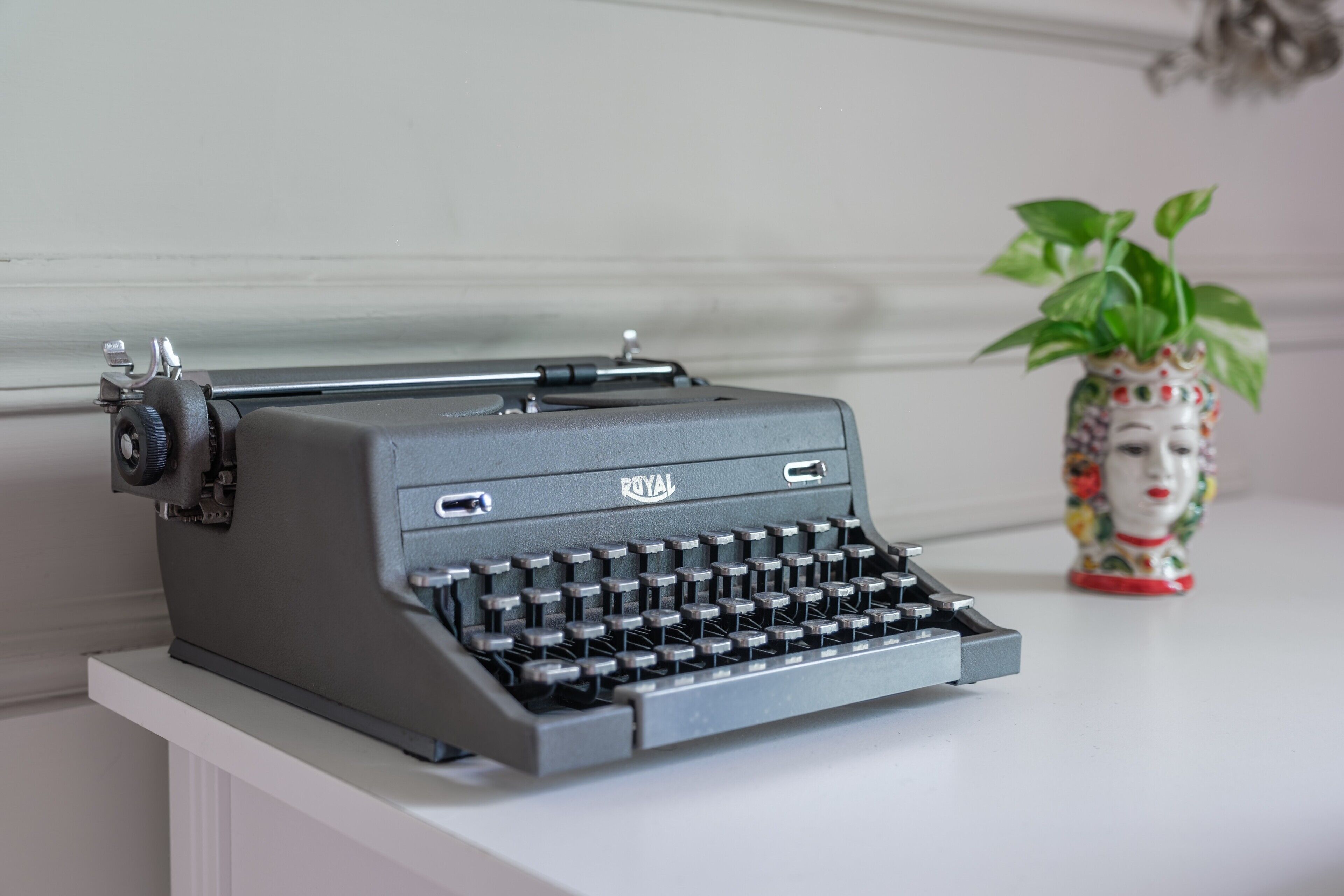 A vintage Royal typewriter on a white surface with a decorative vase and plant in the background.