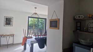 A dining area with a table and chairs, a hanging light fixture, a framed picture on the wall, and a window with a view of greenery.