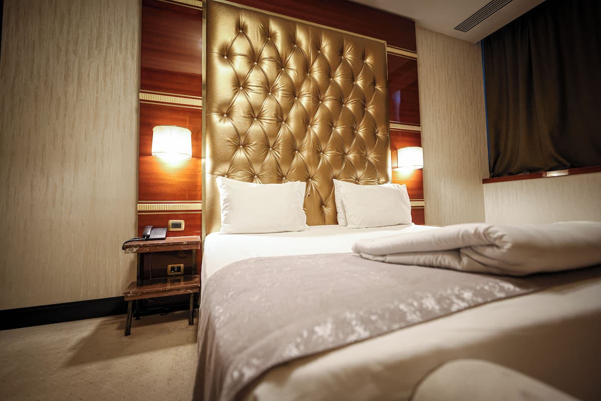 Een hotelkamer met een gestoffeerd hoofdeinde, een nachtkastje met een telefoon, en een net opgemaakt bed met kussens.