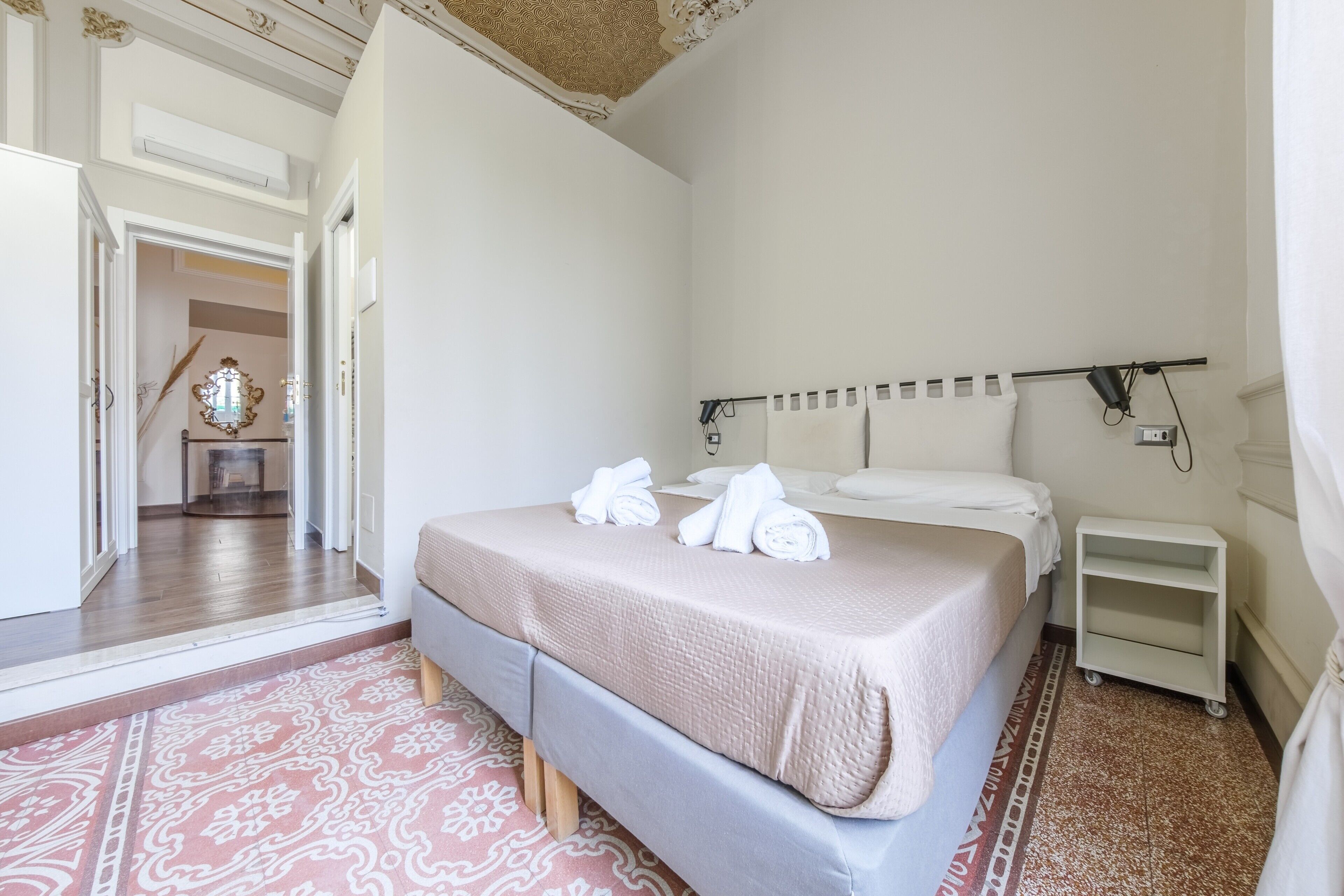 A hotel room with a bed, a bedside table, a wall-mounted lamp, and a small white shelf.