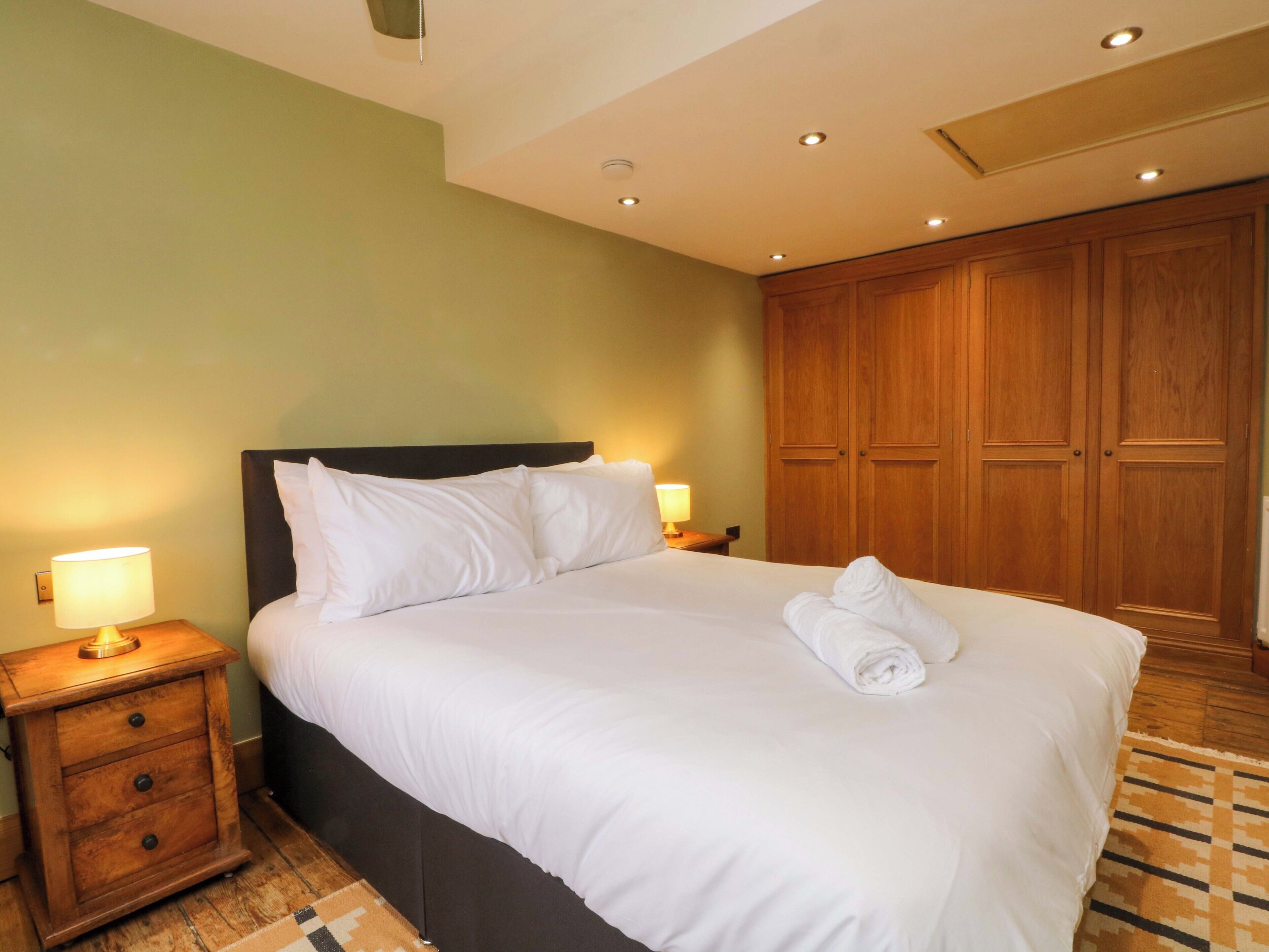 Un lit bien fait, avec du linge de lit blanc, une table de chevet en bois avec une lampe, et une grande armoire en bois dans une chambre d’hôtel.