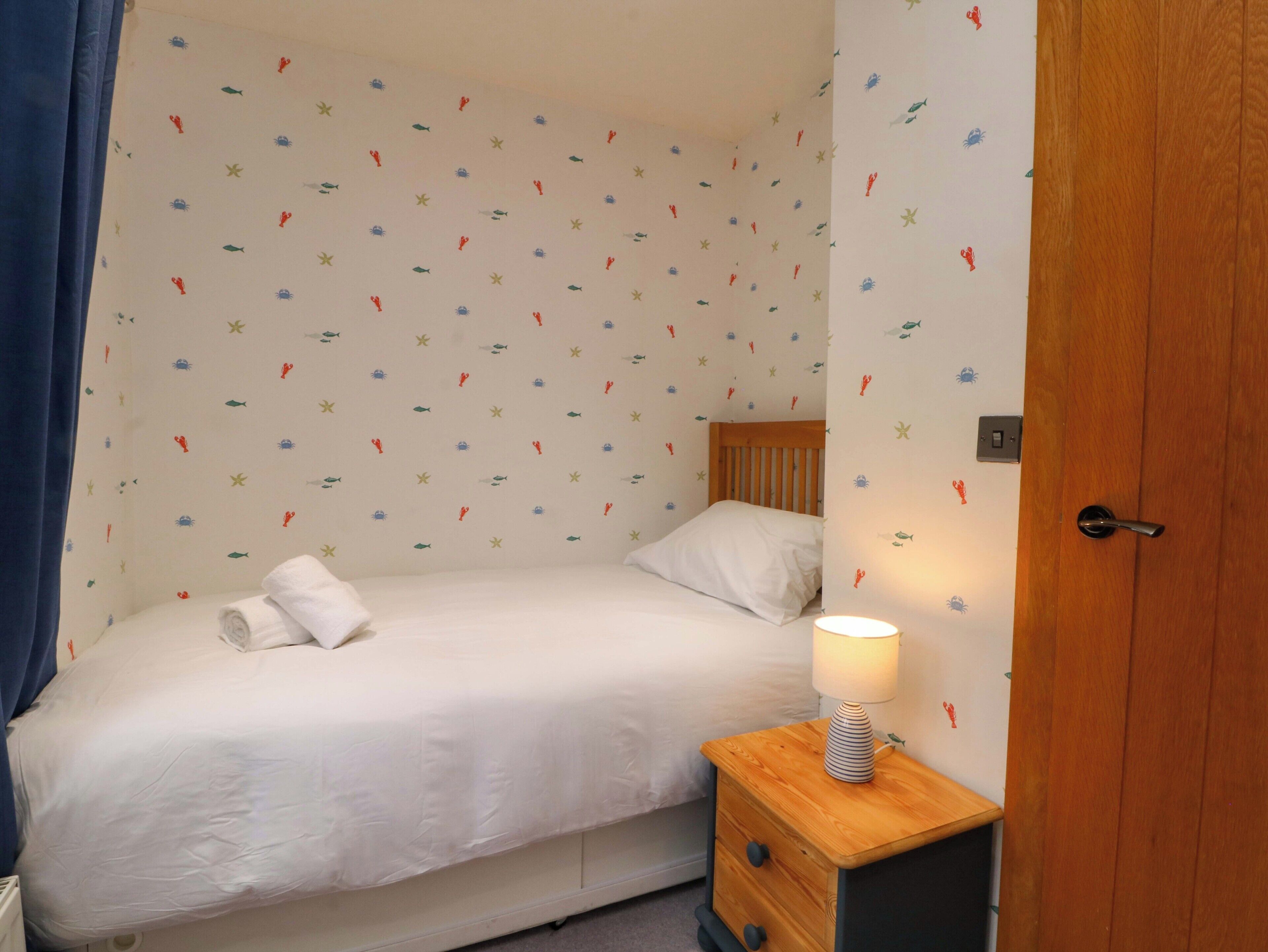 Une petite chambre avec un lit simple, une table de chevet en bois, une couvre-lit blanche et un mur orné d’un motif sur le thème de la marine.