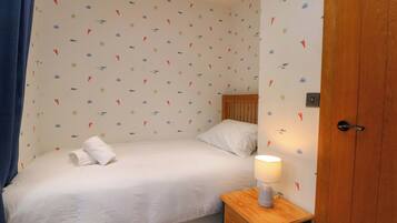 Une petite chambre avec un lit simple, une table de chevet en bois, une couvre-lit blanche et un mur orné d’un motif sur le thème de la marine.