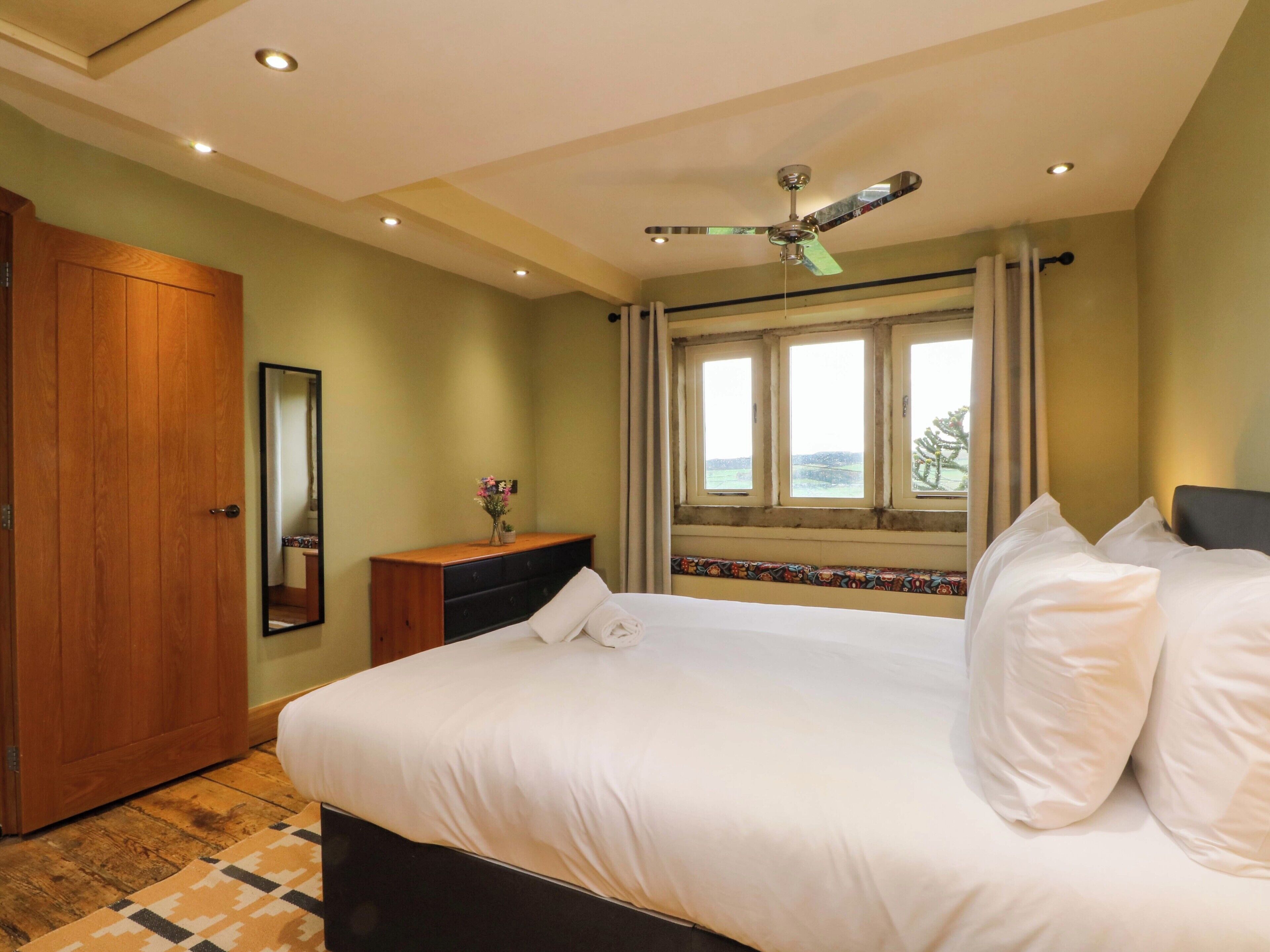 Une chambre à coucher comprenant un lit, une armoire en bois, une commode, un miroir, un ventilateur de plafond et une fenêtre donnant sur un beau paysage.