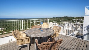 A wooden deck with wicker chairs and a table, overlooking a coastal landscape.