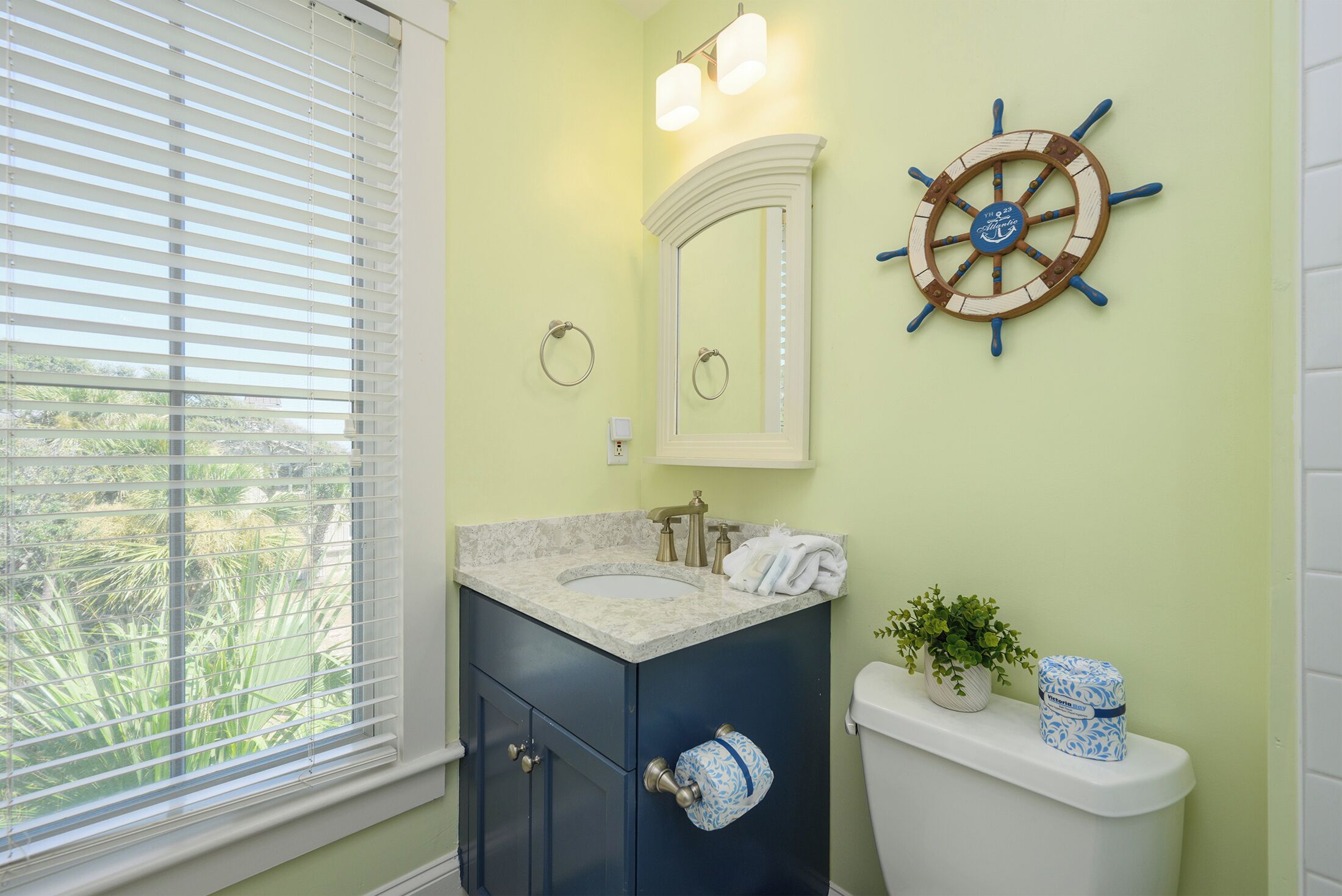 Ein Badezimmer mit Waschbecken, Waschtisch und Toilette. Es gibt ein Fenster mit Jalousien, eine Schiffsschrauben-Dekoration und einen Blumentopf.