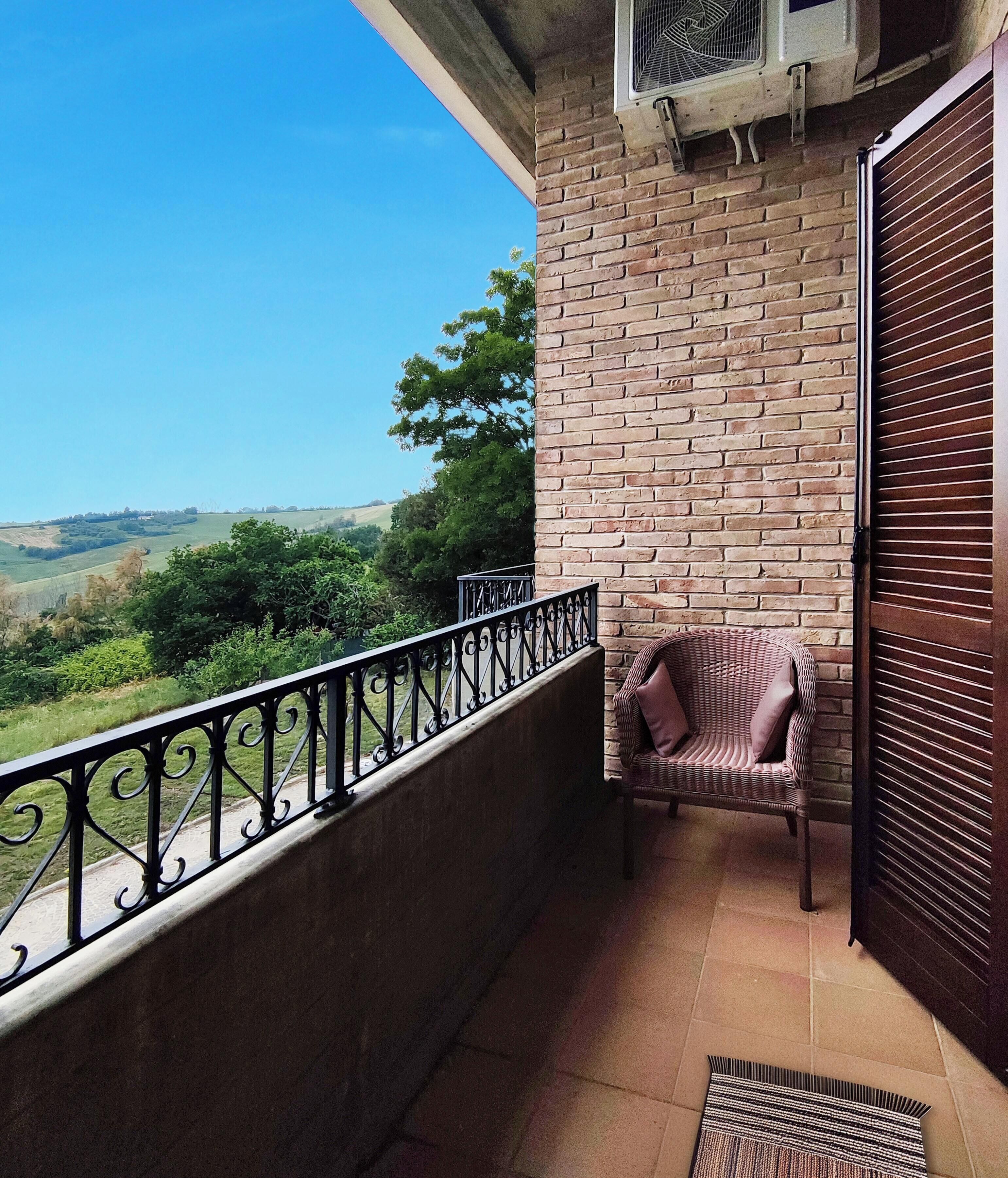 A balcony with a wicker chair, a view of green hills, and a brick wall.