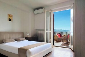 A hotel room with a bed, a nightstand, a wall-mounted air conditioner, and a balcony with wicker furniture.