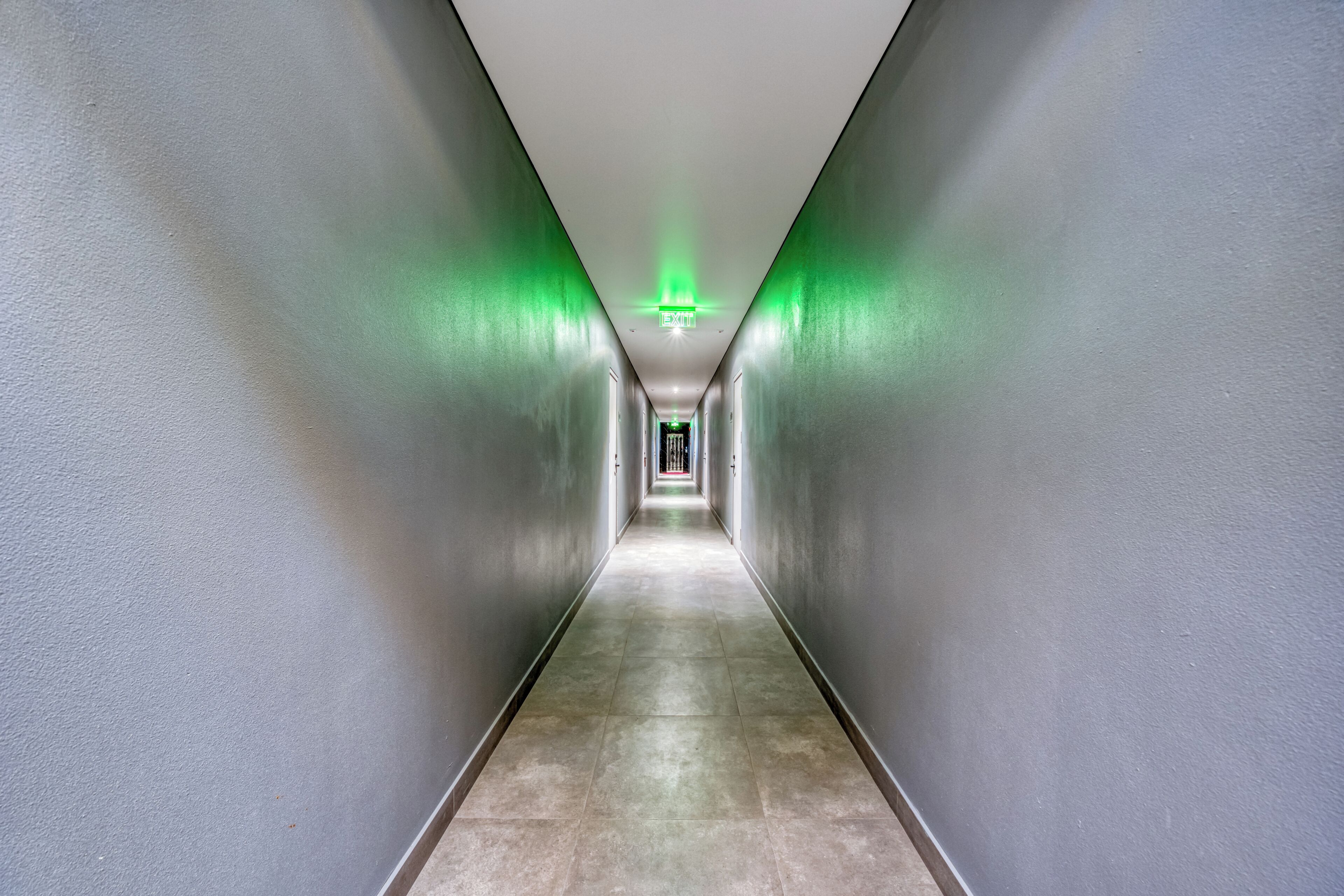 Een lange, smalle gang met een groen vluchtbord aan het einde, begrensd door eenvoudige muren en een betegelde vloer.