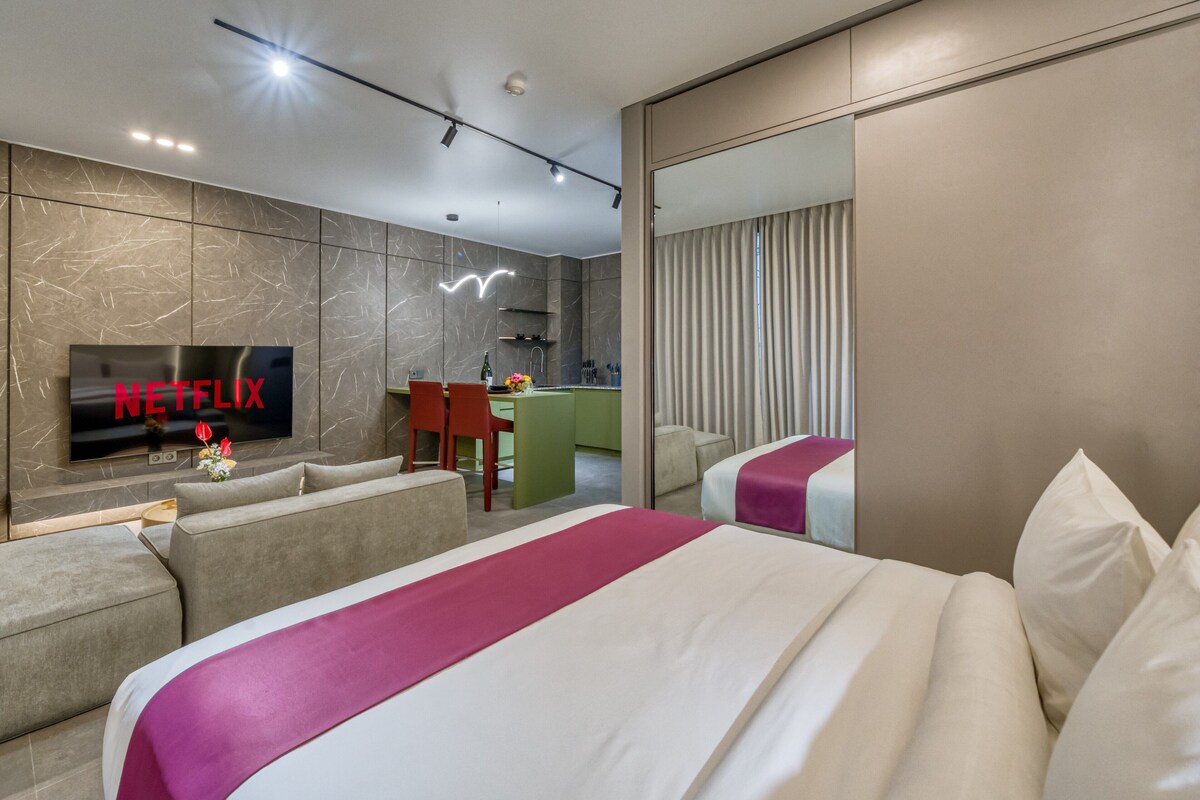 A hotel room with a bed, a sofa, a TV, and a kitchen area.