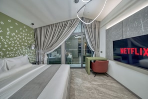Una habitación de hotel moderna con una cama, un televisor mostrando el logotipo de Netflix, una mesita auxiliar verde y un puerta de vidrio que da a un balcón.