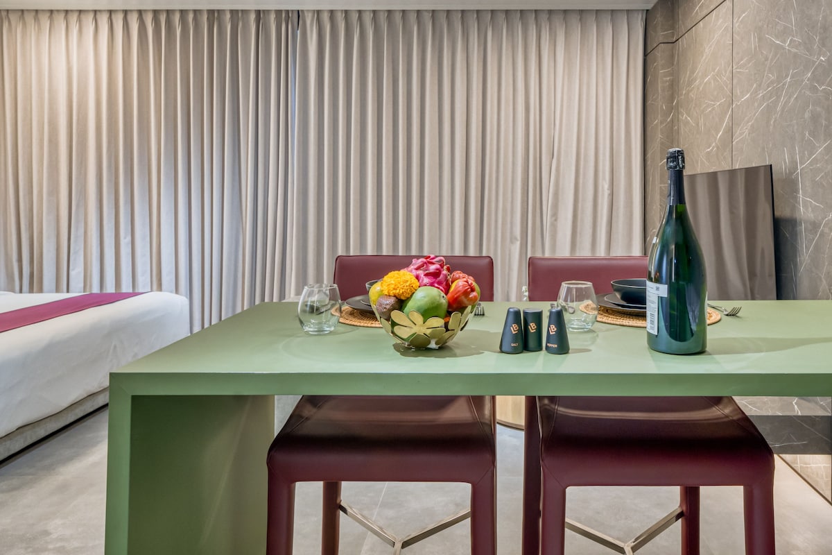A hotel room with a dining table set for two, a bed with a green bedspread, and a curtain backdrop.