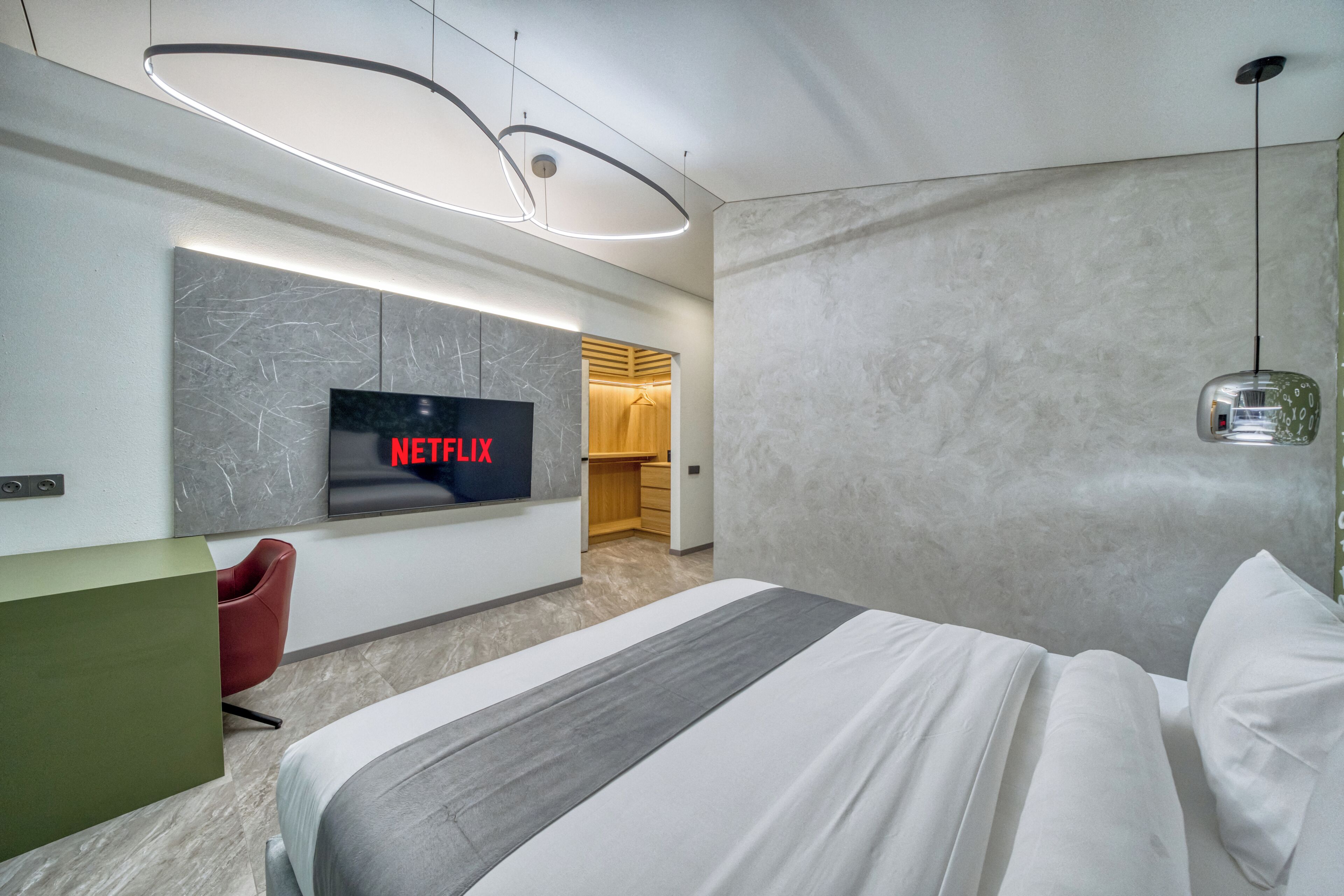 A modern hotel room with a large bed, a TV displaying 'NETFLIX,' a red chair, a green desk, and a wooden wardrobe.
