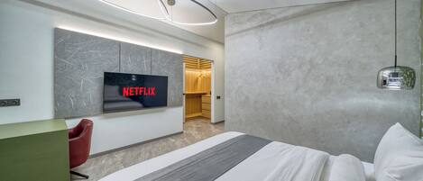 A modern hotel room with a large bed, a TV displaying 'NETFLIX,' a red chair, a green desk, and a wooden wardrobe.