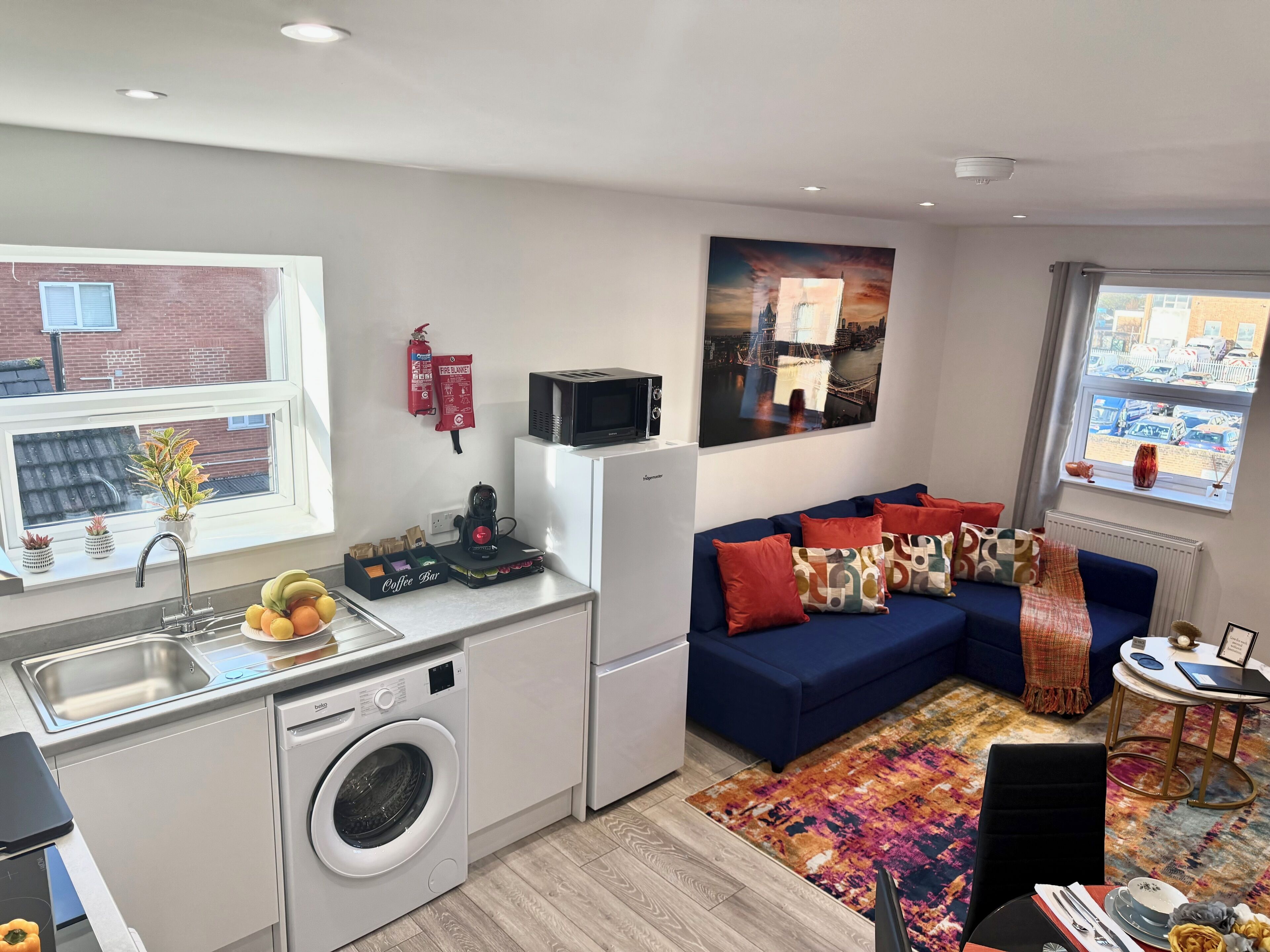 A modern kitchen with a sink, microwave, and coffee machine, a blue sofa with cushions, and a colorful rug.