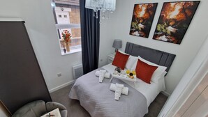 Een verzorgde hotelkamer met een bed, een nachtkastje met een lamp, een stoel met boeken en kunst aan de muren.