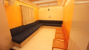 Une pièce avec un canapé de couleur sombre, une petite table et une chaise. Il y a un rideau à la fenêtre et le sol est carrelé.