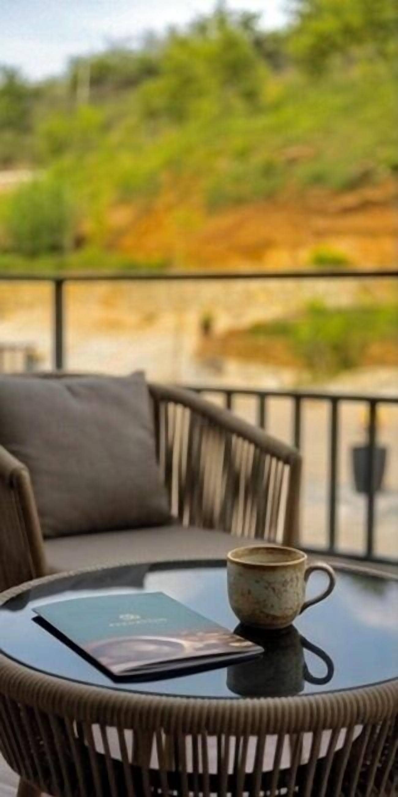 A wicker chair with a cushion, a glass table with a book and a mug, and a view of greenery in the background.