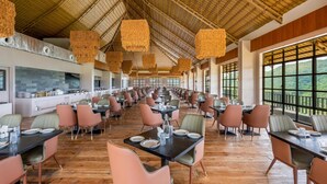 A spacious dining area with high ceilings and large windows offering views of the outdoors. Tables are neatly arranged with chairs, and the room features a modern design with a central kitchen area visible in the background. - Nanda Hill Resort-Aravali Views (Udaipur)