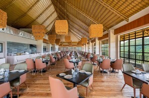 A spacious dining area with high ceilings and large windows offering views of the outdoors. Tables are neatly arranged with chairs, and the room features a modern design with a central kitchen area visible in the background. - Nanda Hill Resort-Aravali Views (Udaipur)