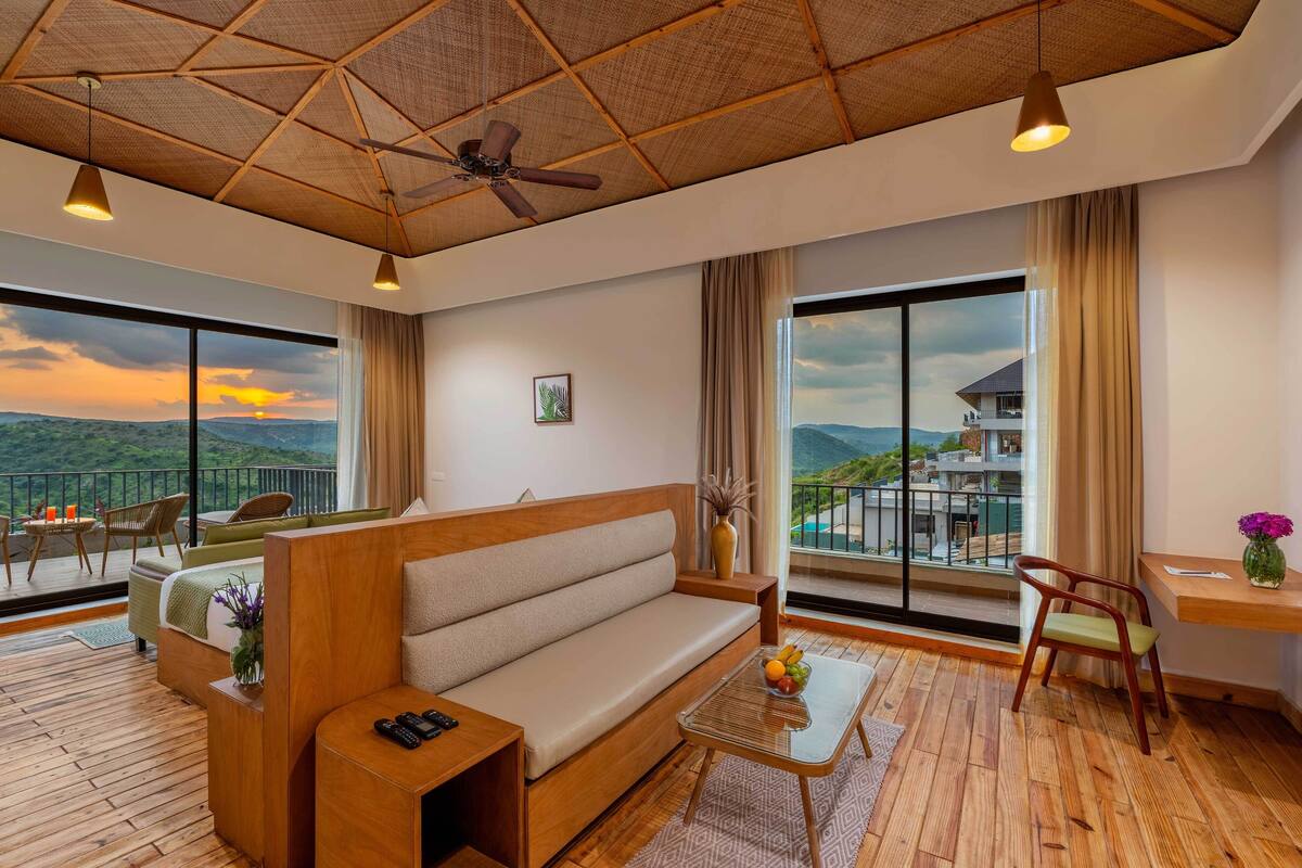 A modern hotel room with a wooden ceiling, large windows offering a view of the mountains, a sofa, a small table, and a chair.