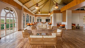A spacious room with a high wooden ceiling, large arched windows, and a central seating area with wicker furniture. - Nanda Hill Resort-Aravali Views (Udaipur)