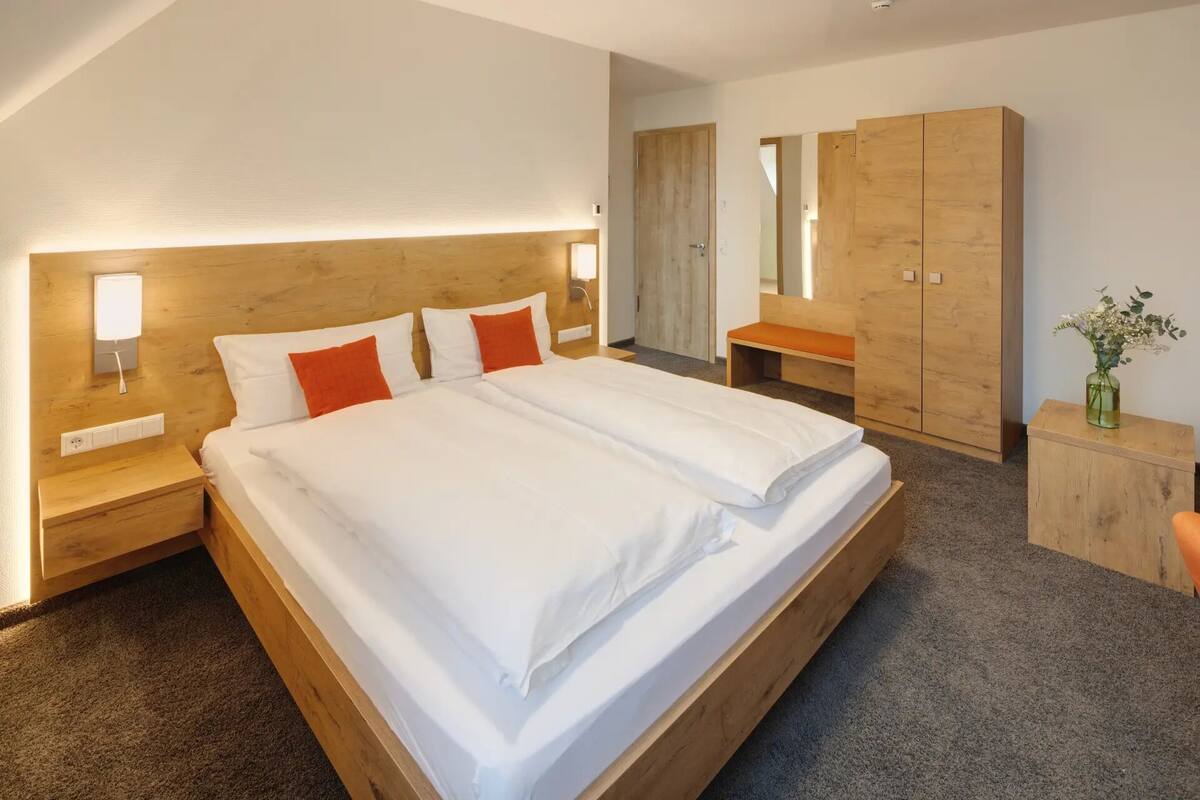 A modern hotel room with a large bed, wooden headboard, bedside lamps, a wooden wardrobe, and a small desk with a vase of flowers.