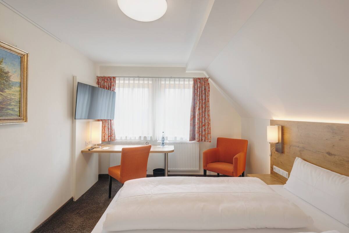 A hotel room with a bed, two orange chairs, a desk with a chair, a television, and a window with curtains.