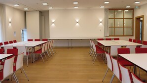 A conference room with white tables and red chairs, equipped with ceiling lights and a large window. - Landhotel Steigenhaus (Untermünkheim)