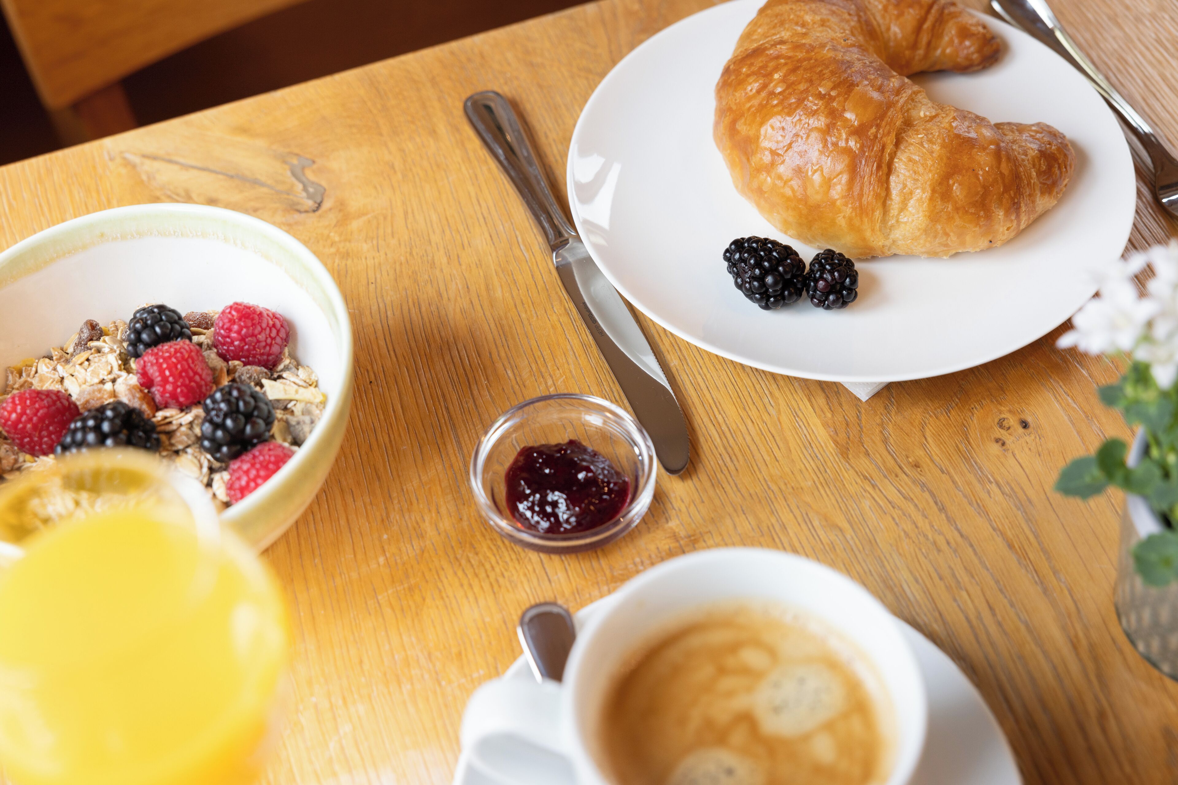 Un desayuno con un croissant, bayas, un tazón de cereal, un recipiente pequeño de mermelada y una taza de café sobre una mesa de madera.